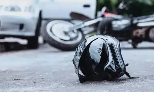 مشهد حادث دراجة نارية يظهر خوذة سوداء على الطريق مع دراجة متضررة وسيارة بيضاء في الخلفية، ما يشير إلى تصادم خطير.