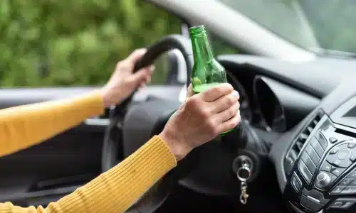 امرأة تقود السيارة وتمسك بعجلة القيادة وبيدها زجاجة خضراء تحتوي على مشروب كحولي.