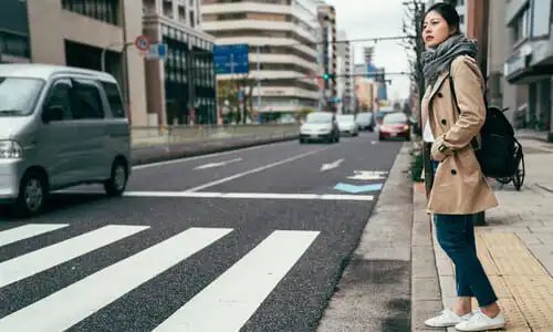 Una mujer asiática a punto de cruzar la calle caminando mientras los coches se acercan por su lado derecho.