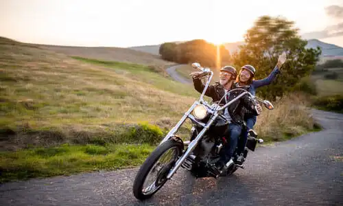 Una pareja que va en una motocicleta por una carretera estrecha en la ladera de una colina a última hora de la tarde.