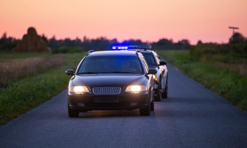 Un auto policial con luces parpadeando acercándose a otro vehículo desde atrás en la calle.