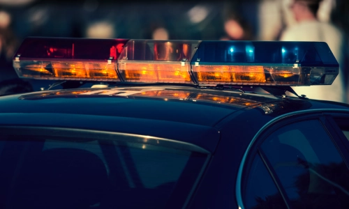 Una captura de cerca de las luces de un auto policial con gente caminando detrás del vehículo en el fondo.