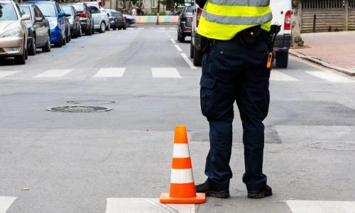 Un oficial de patrulla de pie junto a un cono de tráfico naranja vigilando la escena de un accidente.