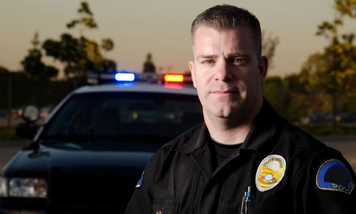 Un oficial de policía de pie junto a su coche y mirando al espectador al atardecer.