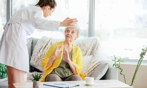 A female caregiver yelling at an old woman seated on a couch in a nursing home.