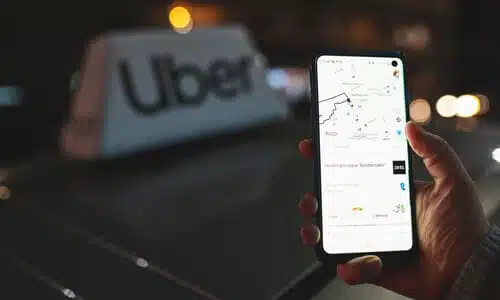 A man's hand holding up a phone displaying a GPS route, with a blurred out Uber vehicle's sign in the background.