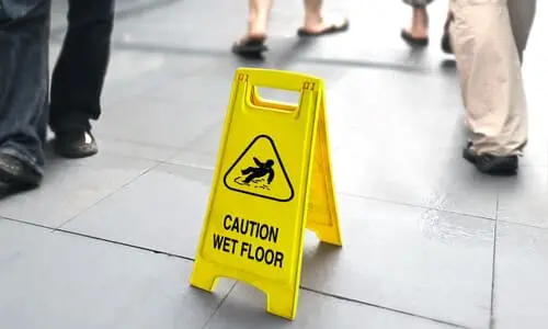 A wet floor sign to prevent slip and fall accidents is pictured next to people walking on a white floor.