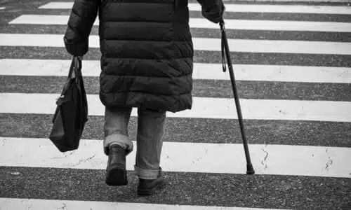 A pedestrian in a jacket and holding a cane as they go along a crosswalk.
