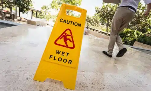 A man losing his balance next to a yellow wet floor warning sign on a walkway.
