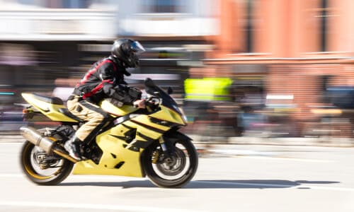 A sportbike speeding through a city street with motion-blurred pedestrians and buildings in the background.