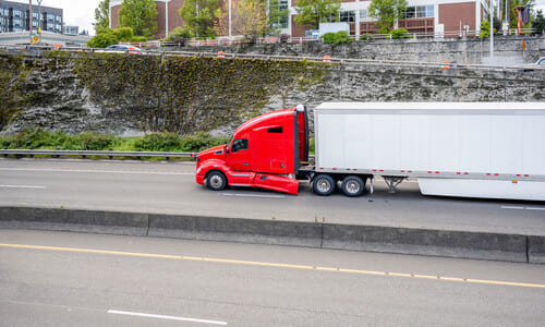 A red semi truck speeding along a highway next to a cutbank with buildings on top of it.