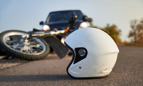 A black motorcycle on its side and with a helmet in the foreground after being struck by a car.