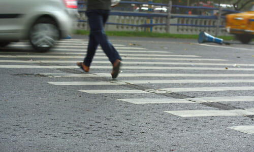 A car zooming past a pedestrian along a crosswalk as they cross the road.