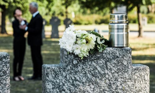 A granite gravestone with a white bouquet on top, with a pair of mourning people in the background.