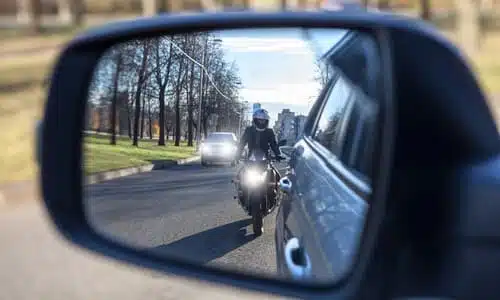 A motorcyclist quickly approaching a car, as seen through a side mirror.