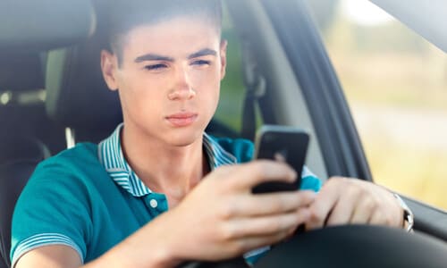 A teenage driver looking at his phone while behind the wheel of his car.