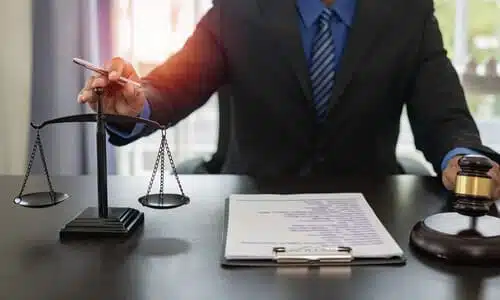 A personal injury lawyer holding his hand over scales and grasping the handle of a gavel while seated at a desk.