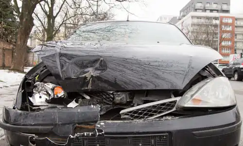 A black car with a busted headlight and crumpled hood after an accident.