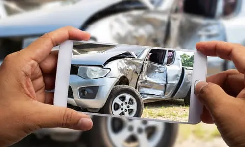 A first-person image of an accident victim raising his phone to document damage to his silver pickup truck.