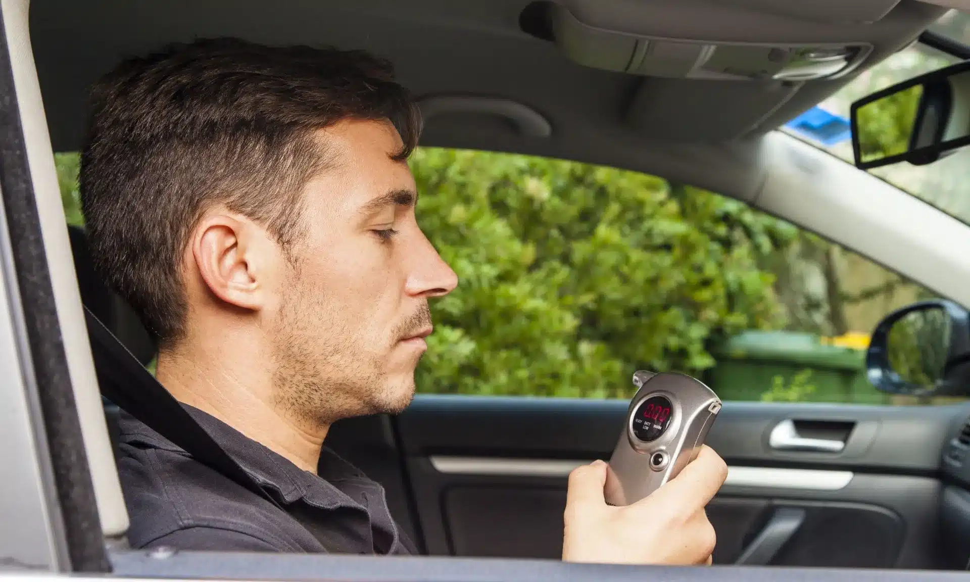 A man in his car staring at a breathalyzer he is about to breathe into.