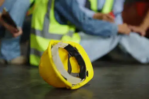 A construction helmet in the foreground with workers helping an injured colleague in the background.