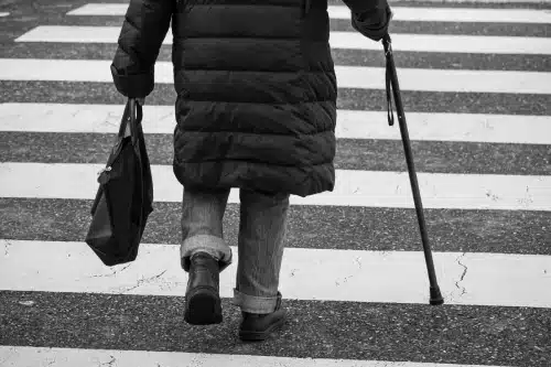A black and white image of a man in thick clothes crossing the road at a pedestrian crossing.