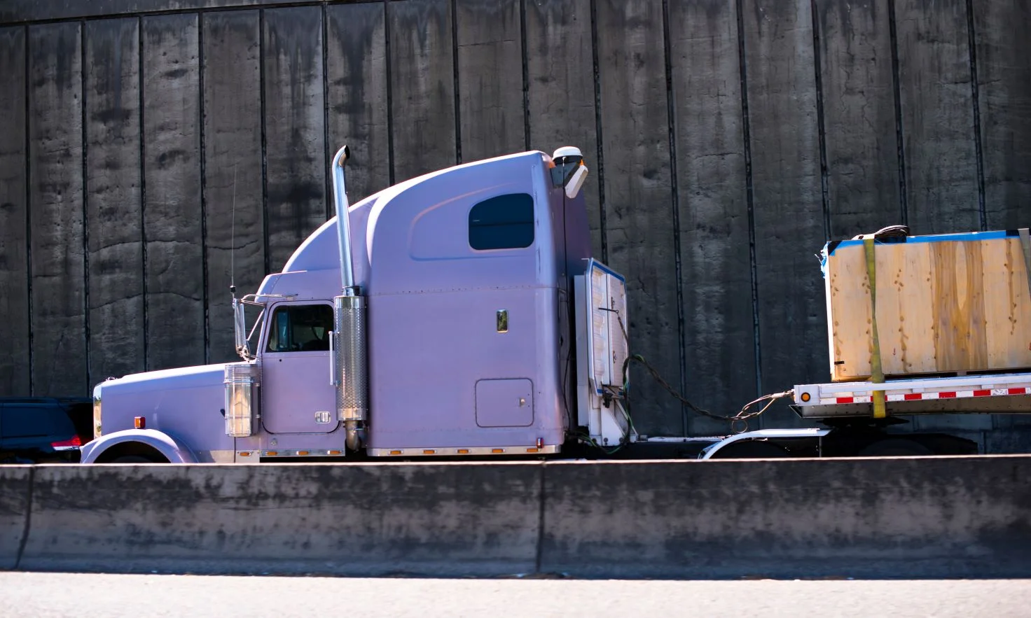 A light-colored semi truck transporting heavy materials driving by a wooden wall.