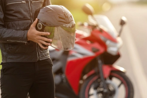 A motorcyclist near his bike holding his helmet next to a rural road.