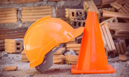 A construction worker's helmet leaning on a safety cone and with construction materials in the background.