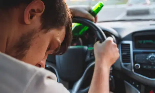 A drunk driver asleep behind the wheel and holding on to a bottle of alcohol.