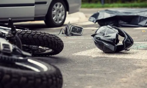 A black motorcycle lying on the road next to a helmet after colliding with a grey car.