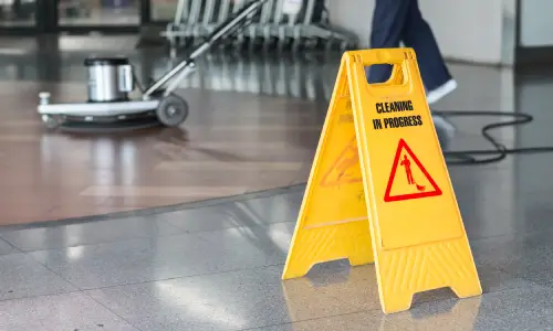 A wet floor sign standing while a maintenance worker polishes the floor in the background.