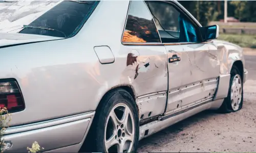 A white car with chipped paint, a broken window, dented door, and flat tire after an accident.