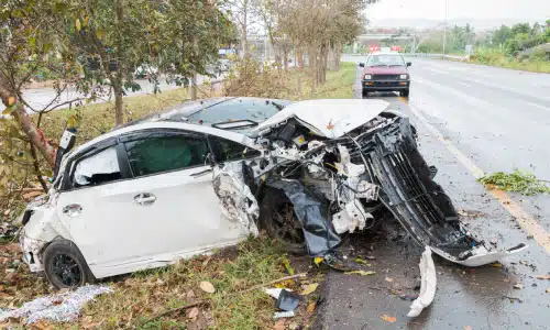 A totaled white car in a ditch after being ran off road by an uninsured driver.