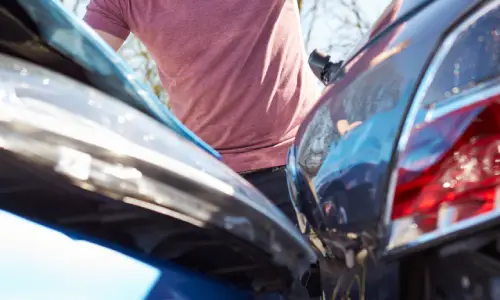 A man in a pink shirt looking at damage from a collision between his car and an uninsured vehicle.