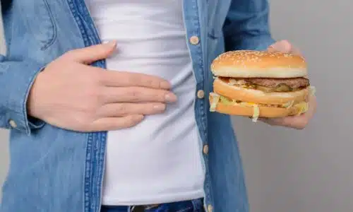 A torso shot of a man holding a burger in one hand and with the other held to his aching stomach.