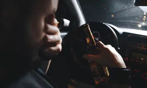 A drunk driver rubbing his eyes and holding a bottle while behind the wheel of a car.