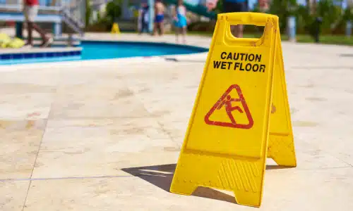 A wet floor sign at the side of a pool on a sunny day.