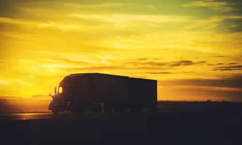 The silhouette of a large semi truck driving on a highway during sunset.