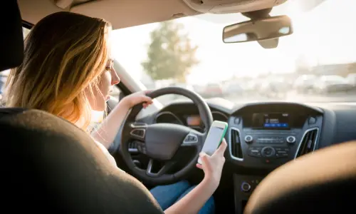 A distracted driver looking at her phone in her right hand while she drives in the afternoon.