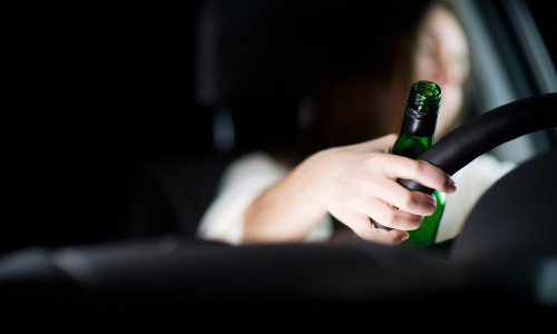 A drunk driver with a bottle in her hand as she operates her vehicle.