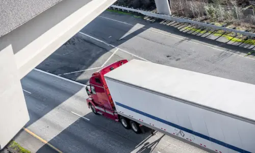 A red semi truck pulling a white container about to drive under a bridge.
