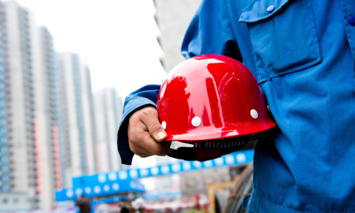 A chest-level shot of a hardhat worker holding a red helmet under his arm.