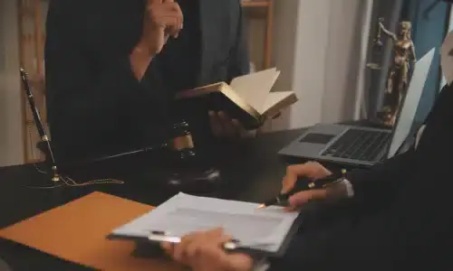 A lawyer reading out of a book while his client fills up forms at the desk.