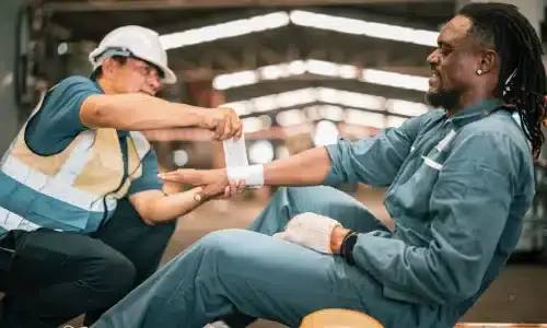 A hardhat worker in a warehouse helping a coworker with an injured arm.