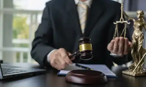 A lawyer at a desk supporting one pan of a scale with one hand and holding a gavel in the other.