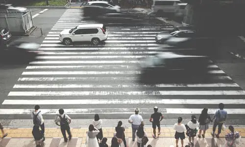 Pedestrians waiting to cross a busy city street at a crosswalk while motion-blurred vehicles zoom by.