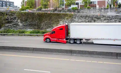 A red semi truck speeding along a highway next to a cutbank with buildings on top of it.