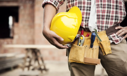 A construction worker with one hand on his waist and the other carrying a yellow hardhat.