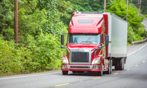A red semi truck driving down a highway in a forest road.